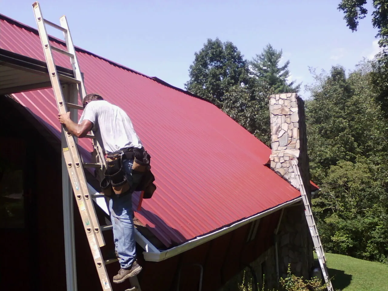 Roofer on a ladder beside a red metal roof and stone chimney — Soffit & Fascia Repair by Roofing Brothers in Varysburg